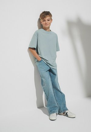 Noor poiss taskus käed, kannab üle mõõdu helerohelist t-särki, laiade säärega teksaseid ja valgeid tossusid, seisab lihtsa valge seina taustal.