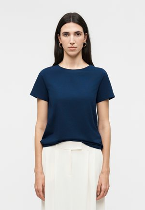 Kobieta z długimi ciemnymi włosami, nosząca złote kolczyki koła, granatową koszulę z krótkim rękawem oraz białe spodnie z wysokim stanem i plisami, stojąca wyprostowana.