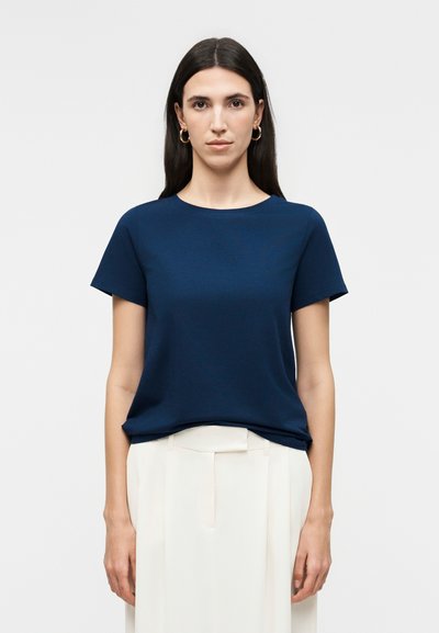 Femme aux longs cheveux foncés portant des boucles d'oreilles créoles dorées, une chemise à manches courtes bleu marine, et un pantalon blanc plissé taille haute, debout droit.