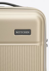 Złota twarda walizka z pionowym żeberkowaniem, czarną etykietą z logo i górnym uchwytem do łatwego przenoszenia.