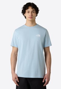 Camiseta de algodón azul claro con mangas cortas y cuello redondo. Presenta un pequeño logo blanco en el lado izquierdo del pecho. Textura suave y lisa.