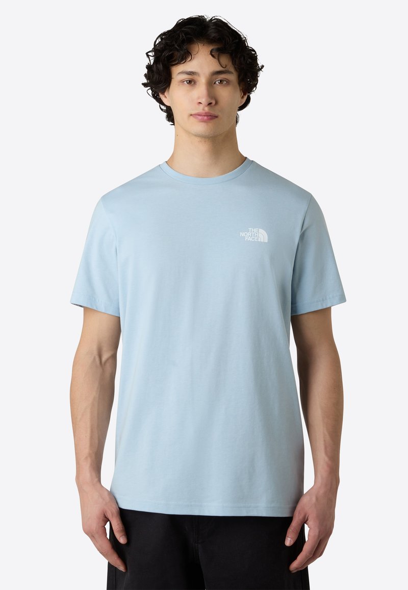 Camiseta de algodón azul claro con mangas cortas y cuello redondo. Presenta un pequeño logo blanco en el lado izquierdo del pecho. Textura suave y lisa.