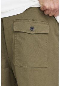 Olivengrønne bukser med et tekstureret stof og en baglomme, der lukkes med en sort knap. Designet inkluderer en klap over lommen.