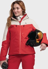 Red and cream ski jacket with zippered pockets, high collar, and attached hood. Glossy black ski helmet with reflective visor held in one hand.