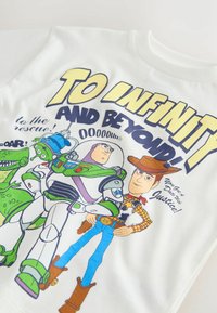 Chemise blanche avec les personnages de Toy Story Woody, Buzz l'Éclair, Rex et un alien vert, avec le texte « Vers l'infini et au-delà ! » en lettres grasses.