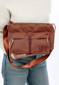 Borsa a tracolla in rete di bronzo con due tasche anteriori con zip, tracolla regolabile e testo in rilievo. Esterno testurizzato e dettagli hardware lisci.