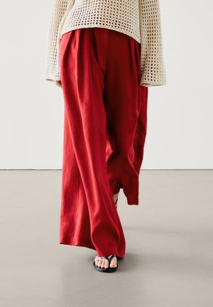 Personne marchant portant un pantalon rouge ample, un pull en tricot crème et des sandales noires à bout ouvert sur un sol uni et un fond blanc.