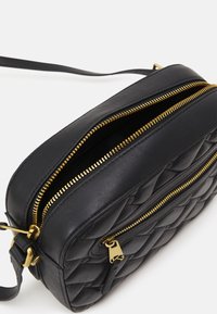 Črna usnjena torba za čez ramo z ukrivljenimi vzorci, dvema zlatima zadrgama in snemljivim trakom. Gladka tekstura, srednja pravokotna oblika.