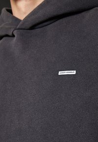 Σκούρο γκρι φούτερ με απαλή υφή, διαθέτει κουκούλα και μια λευκή κεντημένη ετικέτα "UNDER ARMOUR" στο μπροστινό μέρος.