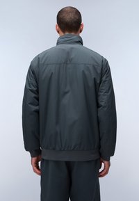 Giacca a vento grigia con collo alto, polsini e vita elasticizzati; texture liscia con un leggero lucido. Design minimalista, senza loghi visibili.
