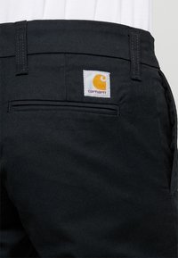 Zwarte broek met een achterzak en een vierkant, wit label met een oranje logo. Gelaagd materiaal met een tijdloos ontwerp.