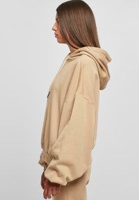 Beige oversized kapucnis hoodie, zsinóros kivitelben, bordázott mandzsettákkal és laza szabással; az oldalról látható puha textúra és laza dizájn.