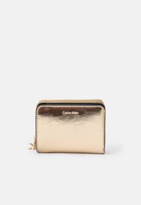 Portefeuille en métal doré avec une texture lisse, fermeture à glissière et logo de la marque en relief. Présente une forme rectangulaire compacte et des finitions en doré.