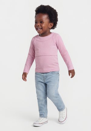 Mosolygós, göndör hajú kisgyermek világos rózsaszín hosszú ujjú pólóban, világoskék farmerben és fehér tornacipőben, előre sétál.