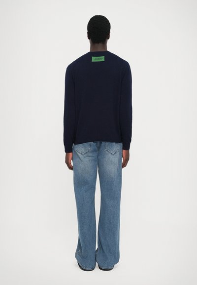 Pulover bleumarin cu decolteu rotund, mâneci lungi și etichetă verde pe spate, asortat cu blugi drepti din denim de culoare albastru deschis.