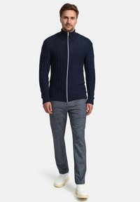 Marineblauer, gerippter Reißverschluss-Pullover mit hohem Kragen, kombiniert mit strukturierten grauen Hosen und weißen Sneakers, die einen abgestimmten, legeren Look erzeugen.