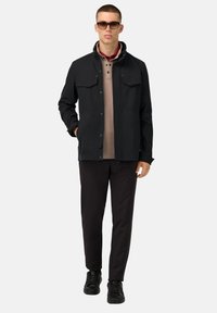 Giacca nera con bottoni, dotata di due tasche sul petto, indossata sopra una maglietta polo color beige. Pantaloni neri slim e sneakers nere completano l'outfit.