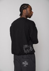 Mężczyzna z plecionymi włosami, ubrany w czarną, szeroką koszulę i spodnie z logo "Multiply Apparel", zwrócony bokiem na jednolitym tle.