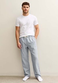 Witte T-shirt gecombineerd met lichtblauw gestreepte pyjamabroek. De broek heeft een elastische tailleband met een trekkoord. Model staat blootsvoets.