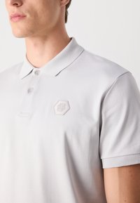 Polo de cor cinza claro com uma textura suave, golas de botão duplo e um discreto logótipo hexagonal no lado esquerdo do peito. Manga curta.
