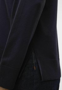 Pull en coton bleu marine avec des poignets côtelés et des fentes latérales, présentant une texture lisse et un design décontracté. Denim visible en dessous.