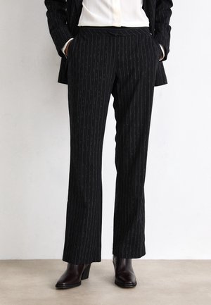 Osoba nosząca czarne spodnie w prążki, ciemne buty na obcasie, białą koszulę oraz dopasowaną czarną marynarkę w prążki, ręce w kieszeniach.