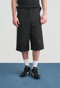 Άνδρας που φοράει μαύρα μακριά σορτς με λευκές πλευρικές ρίγες, λευκές κάλτσες μέχρι τον αστράγαλο και μαύρα παπούτσια Puma, στέκεται σε μπλε χαλί.