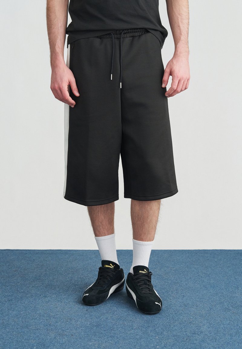 Άνδρας που φοράει μαύρα μακριά σορτς με λευκές πλευρικές ρίγες, λευκές κάλτσες μέχρι τον αστράγαλο και μαύρα παπούτσια Puma, στέκεται σε μπλε χαλί.