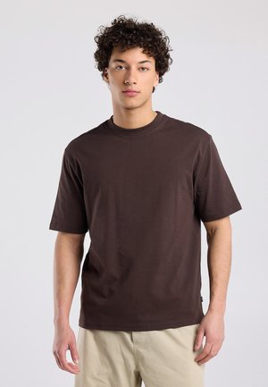 Jonge man met krullend haar, gekleed in een effen donkerbruine korte mouw T-shirt en licht beige broek, staand tegen een lichte achtergrond.