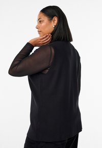 Vrouw met steil haar op schouderlengte, die een mouwloze zwarte vest draagt over een doorschijnende zwarte top met lange mouwen, en met haar rechterhand haar nek aanraakt.