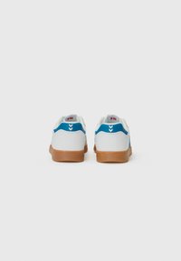 White sneakers with a blue accent, featuring a textured gum rubber sole, and a minimalist design with chevron detailing on the heel.