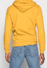 Persona con una sudadera amarilla y jeans de mezclilla azul claros, vista desde atrás con las manos relajadas a los lados.