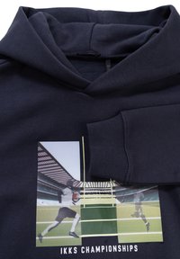 Sudadera azul marino con un estampado gráfico que muestra una escena deportiva. Presenta un bolsillo frontal y puños acanalados. Tejido suave y texturizado.