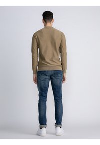 Langärmliger khakifarbener Pullover und blaue Jeans mit einer ausgewaschenen Optik. Das Outfit wird mit weißen Sneakers und einem lockeren Schnitt abgerundet.