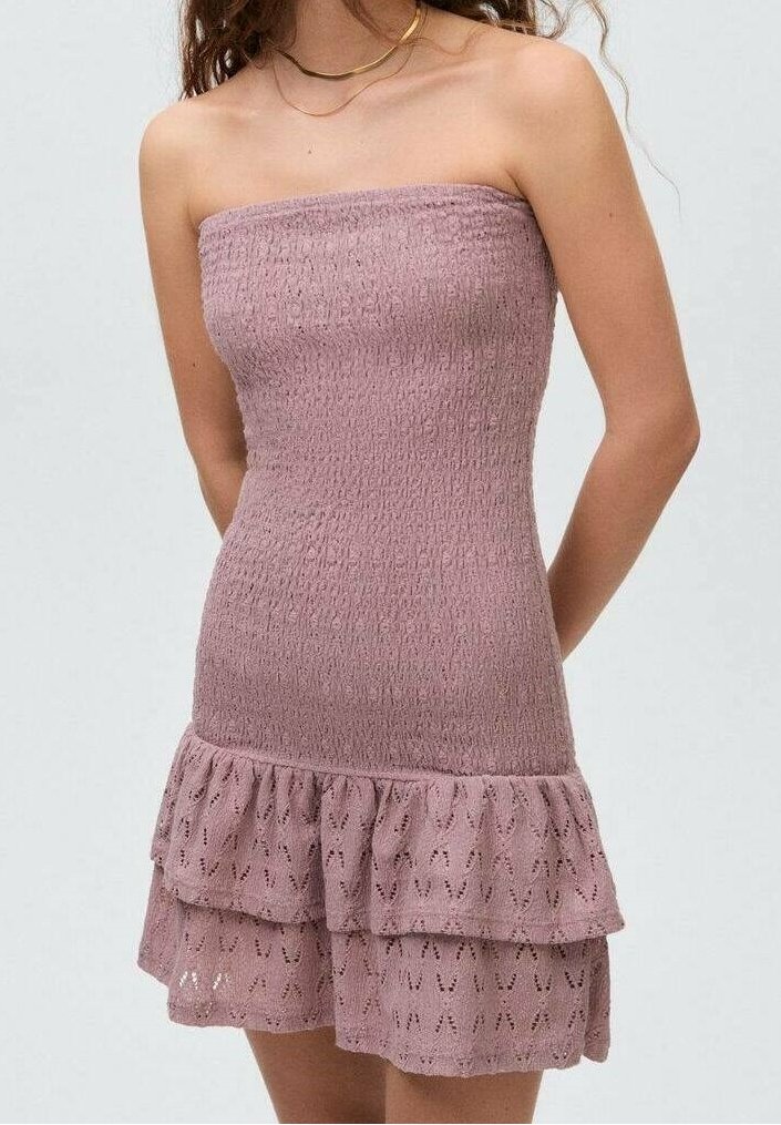 Femme portant une robe mini lilas texturée sans bretelles avec un ourlet à volants superposés et un collier ras de cou doré sur un fond uni.