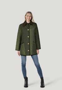 Grün gesteppter Mantel mit goldenen Knöpfen, einem braunen Kragen und zwei Seitentaschen. Getragen über blauen Jeans und schwarzen Stiefeln.