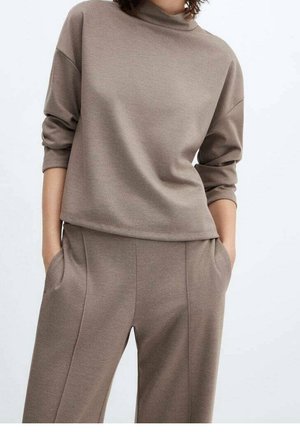 Femme portant un haut taupe à manches longues avec col montant et un pantalon large assorti, mains dans les poches, sur un fond uni.