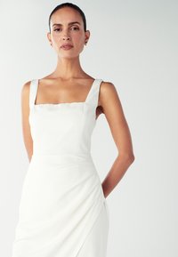 Žena s tamnom kosom nosi bijelu haljinu bez rukava sa širokim trakama i kvadratnim izrezom, stojeći nasuprot jednostavne svijetle pozadine.