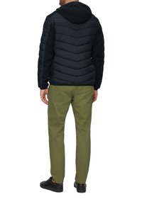 Donkerblauwe pufferjack met een capuchon, voorzien van een chevronpatroon op de achterkant en zwarte mouwen. Olijfgroene broek en zwarte schoenen maken de outfit compleet.