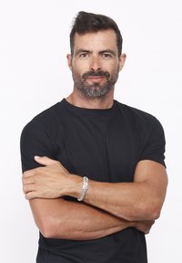 T-shirt nera a girocollo, braccia muscolose incrociate, indossa un bracciale d'argento con design a catena, capelli corti neri e barba. Sfondo bianco.