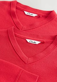 Maglioni rossi a maglia con scollo a V, caratterizzati da polsini a coste e una texture liscia. Le etichette riportano "Clarks". Due maglioni esposti sovrapposti.