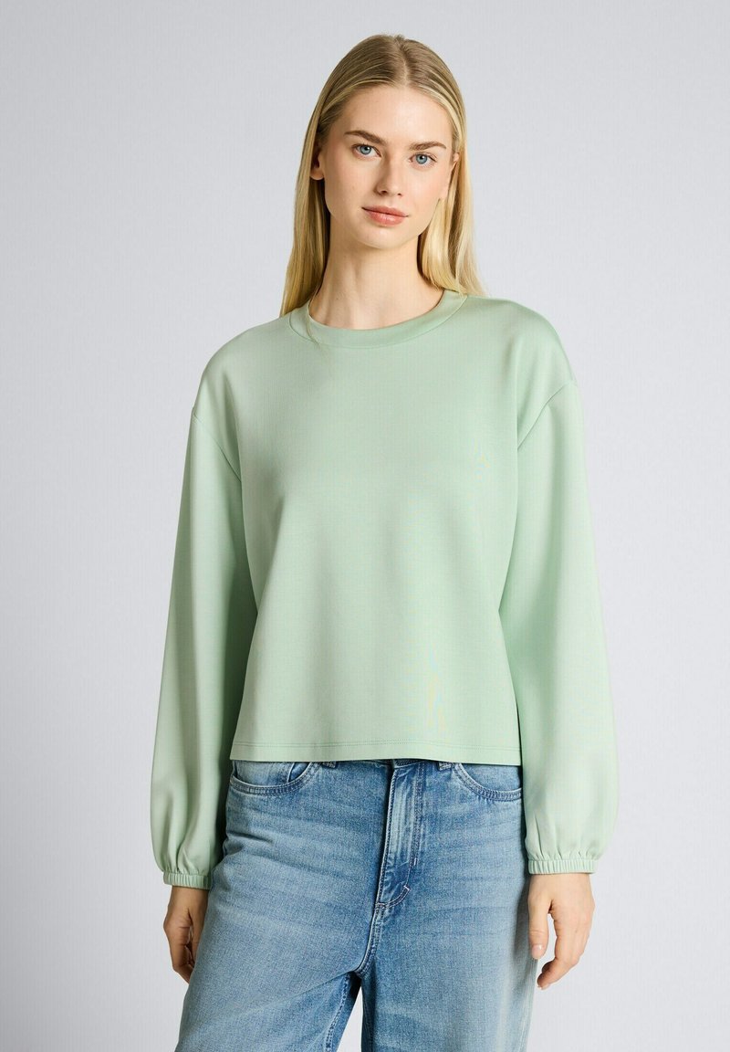 Junge Frau mit langen blonden Haaren trägt ein hellgrünes Langarmshirt und blaue Jeans, steht vor einem schlichten grauen Hintergrund.