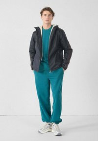 Jonge man staand, gekleed in turquoise joggingbroek en sweatshirt, met een zwart ritsvest met capuchon en witte sneakers, tegen een effen achtergrond.