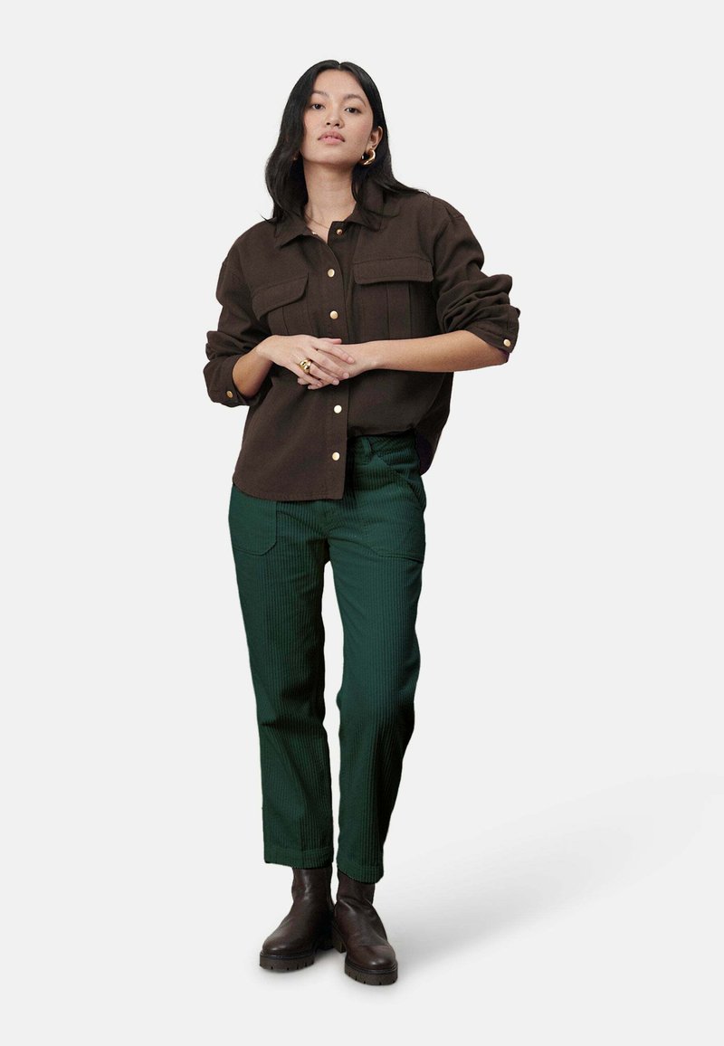 Brown button-up shirt met twee borstzakken, gecombineerd met groene ribgebreide broeken en donkerbruine enkellaarsjes. Model poseert met de handen gekruist.