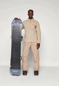 Snowboard en uitrusting: beige ritsfleecejack, bijpassende broek en een zwarte snowboard met kleurrijke grafieken. De laarzen zijn zwart met een robuust ontwerp.