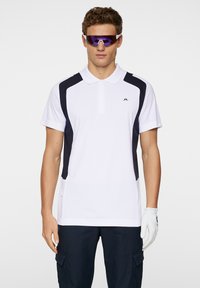 Vit polo skjorta med marinblå accenter, korta ärmar och krage. Har en logotyp på bröstet och en strukturerad design på sidopanelerna.