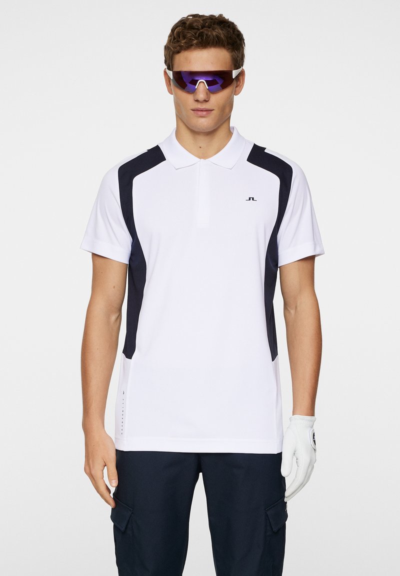 Vit polo skjorta med marinblå accenter, korta ärmar och krage. Har en logotyp på bröstet och en strukturerad design på sidopanelerna.