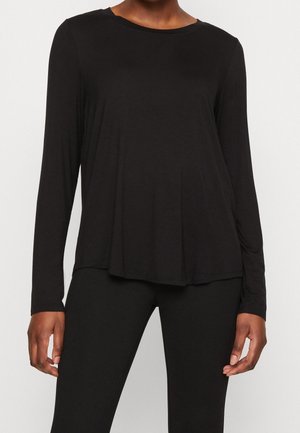 Personne portant un haut noir ajusté à manches longues et un pantalon noir, debout devant un fond clair uni, les bras relâchés le long du corps.