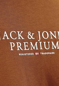 Rudas medžiaga su balta užrašu „JACK & JONES PREMIUM“, šalia jo mažas registruotas prekės ženklas ir Danijos vėliavos detalė.