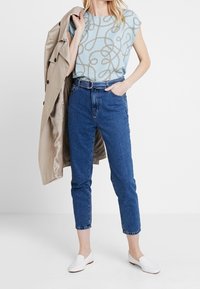 Top de manga corta azul claro con un patrón de lazos, combinado con jeans de talle alto azules y zapatos blancos sin cordones, complementado con un abrigo beige.